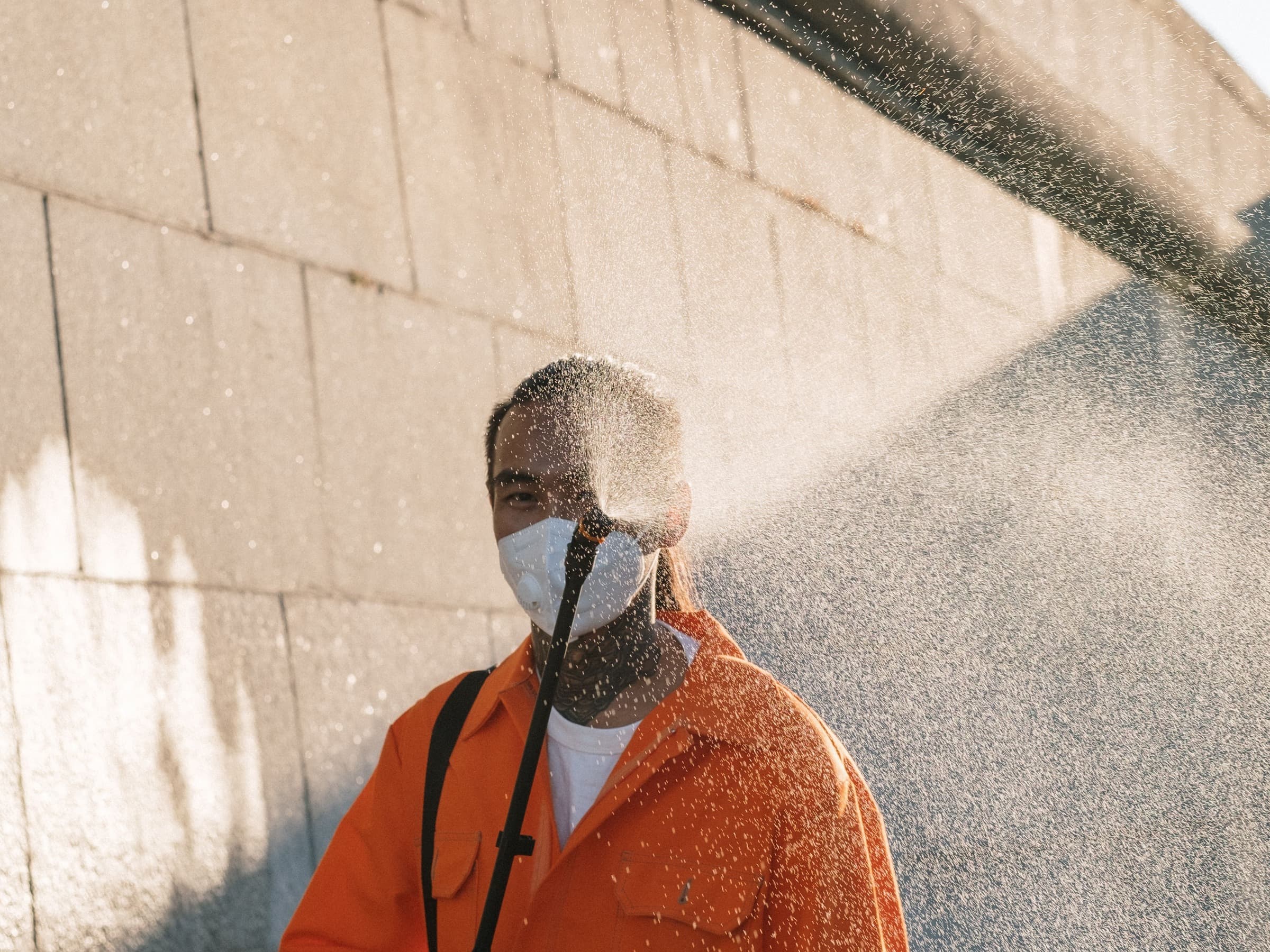 Technician in orange coveralls and a mask washing an exterior wall with a spray wand