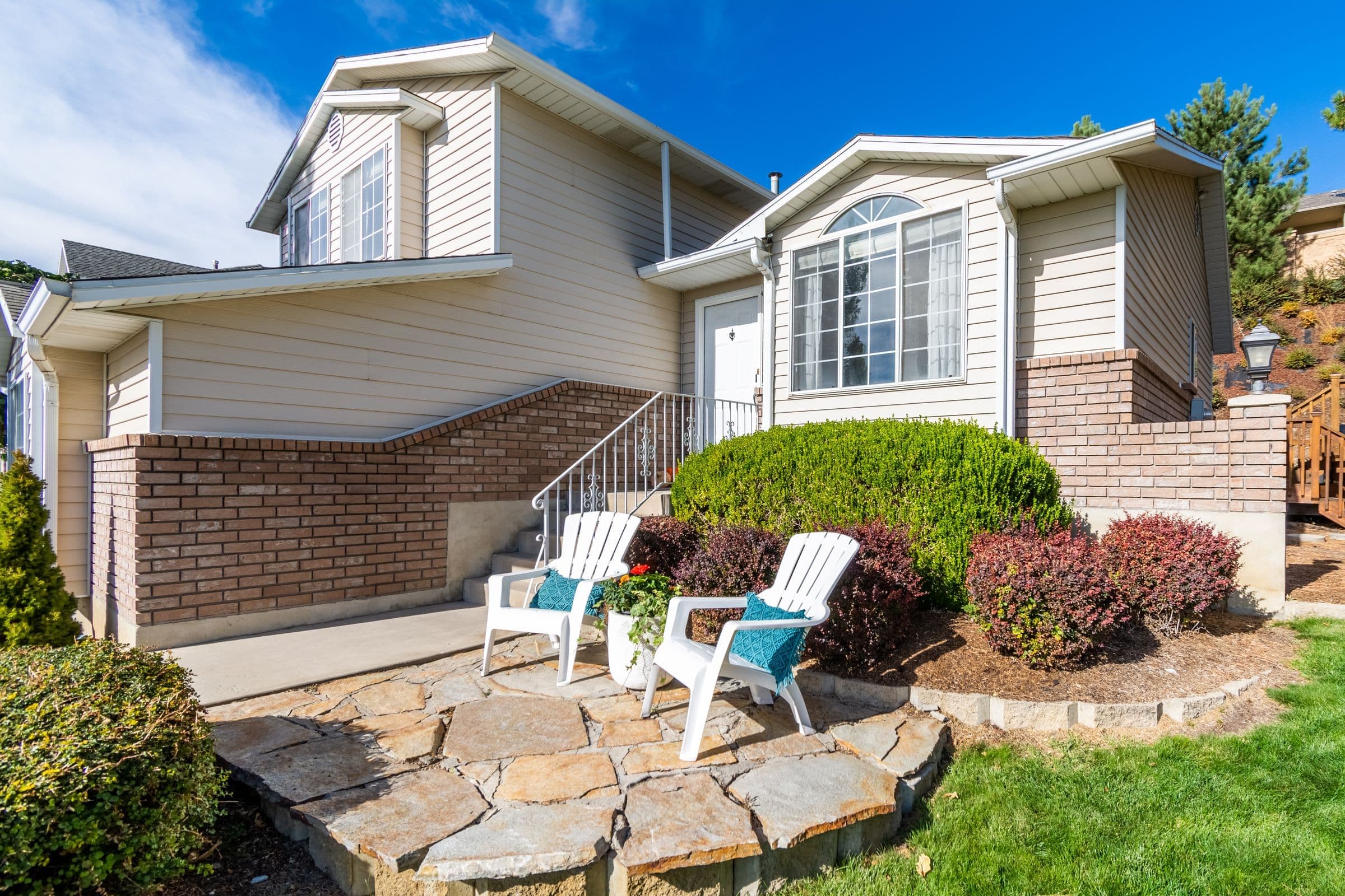 Split-level home with beige siding, a brick base and a stone patio with two Adirondack chairs